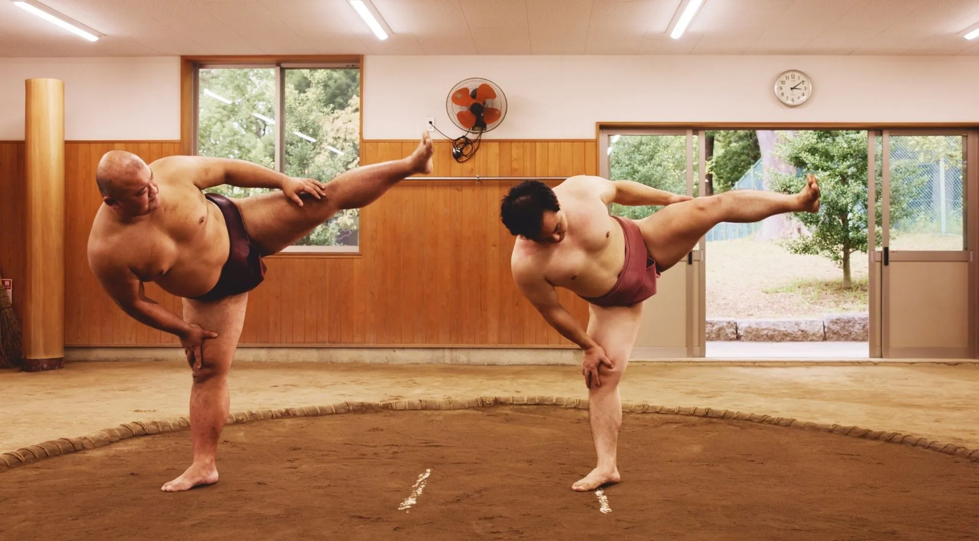 Two wrestlers doing a synchronized leg-stretch shiko exercise in a practice stable