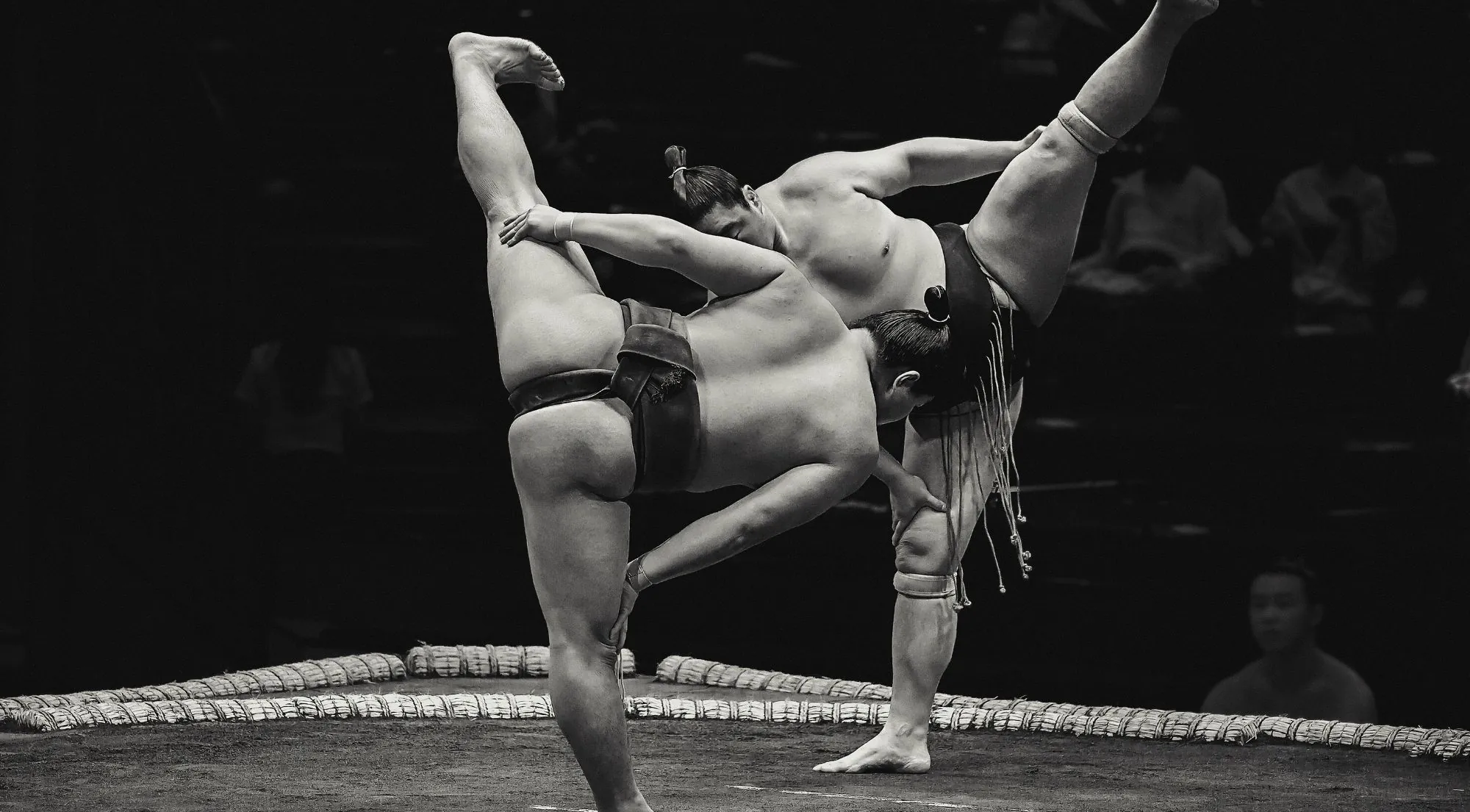 Black and white photo of a wrestler mid-bout raising his leg in the shiko stomp during a live tournament, referee visible