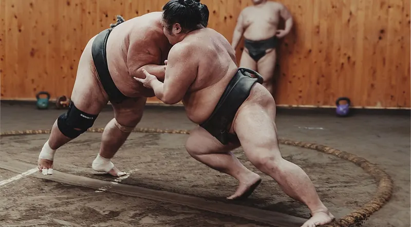 Two wrestlers grappling during practice in a stable, kettlebells visible in the background